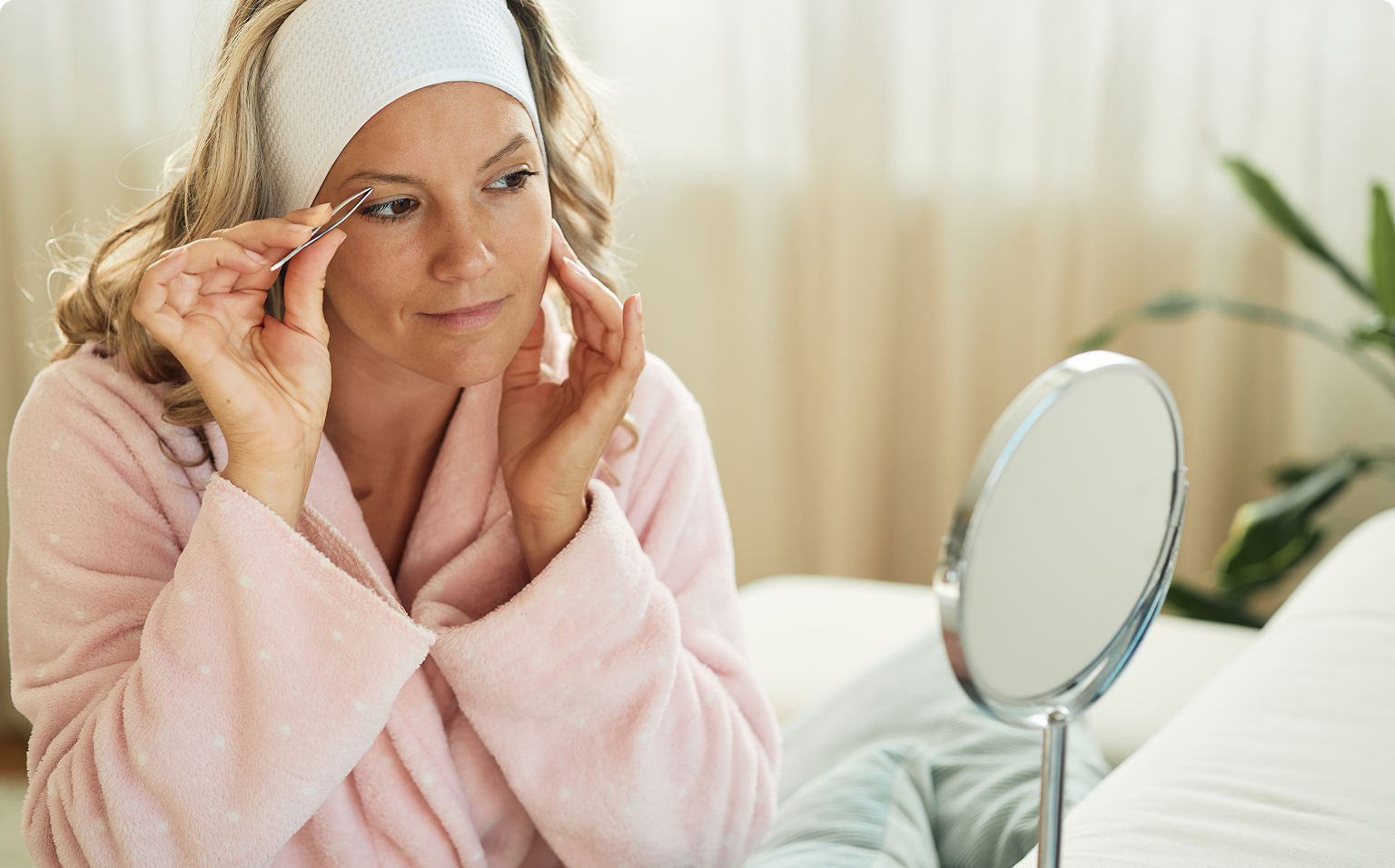 Woman tweezing eyebrows in pink robe.