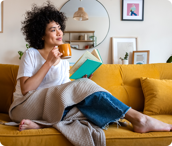Woman reading book, drinking coffee on couch.
